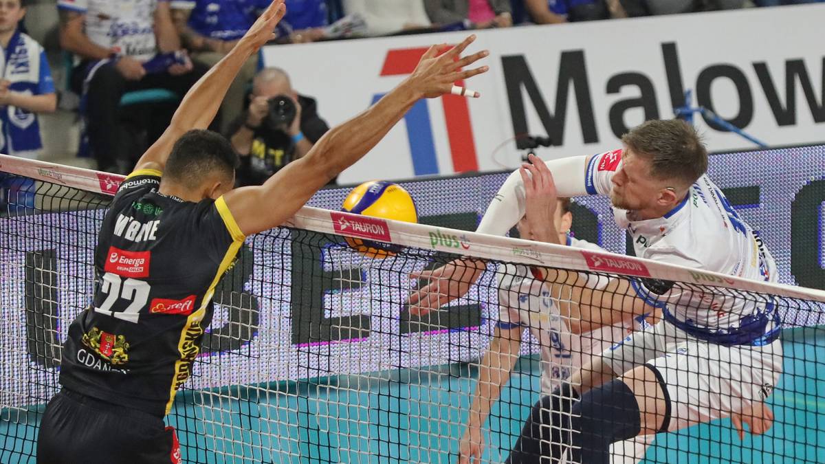 Siatkarz w czarnej koszulce z numerem 22 pr&oacute;buje zablokować atak przeciwnika w białej koszulce przy siatce.
