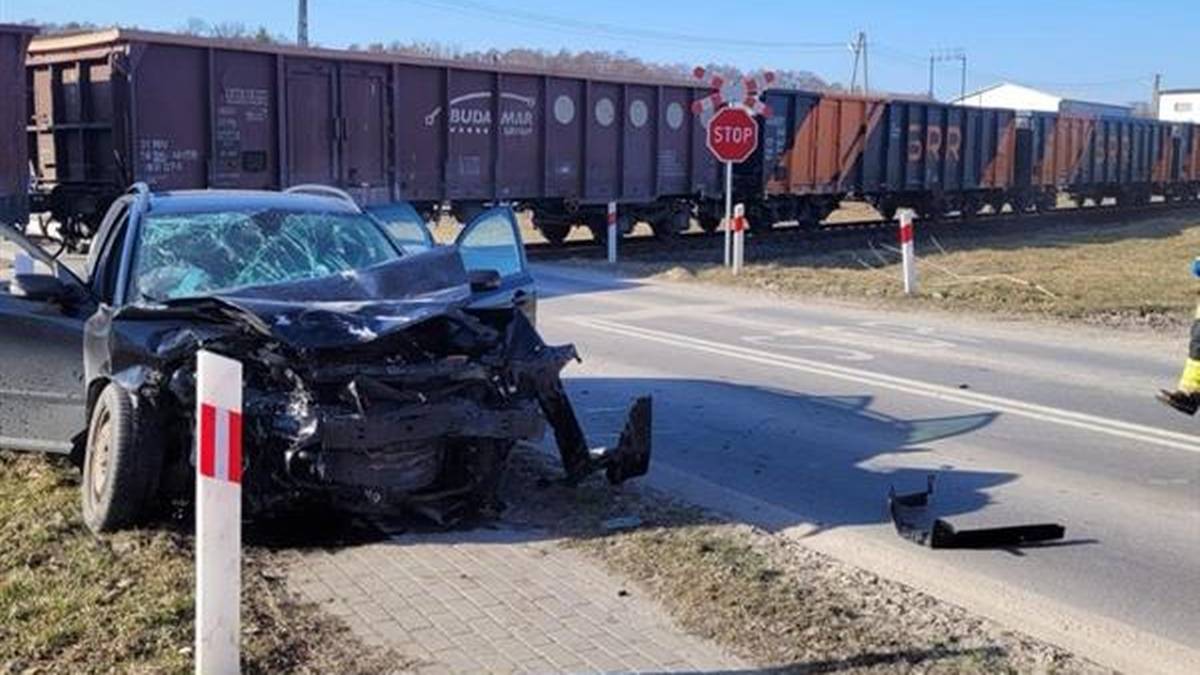 Zniszczony czarny samoch&oacute;d osobowy na poboczu drogi, z otwartymi drzwiami, obok tor&oacute;w kolejowych z długim pociągiem towarowym w tle.