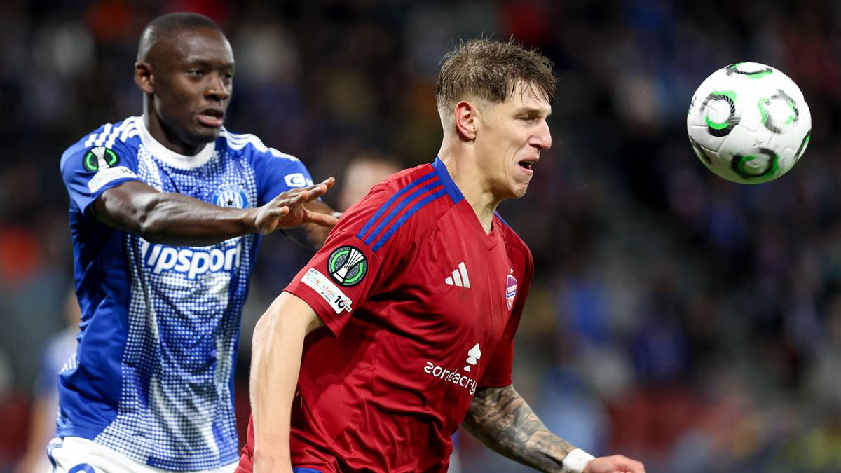 Dwóch piłkarzy walczy o piłkę podczas meczu Ligi Konferencji. Piłkarz w czerwonej koszulce w ruchu, piłkarz w niebieskiej koszulce próbuje go zatrzymać. Piłka w powietrzu.