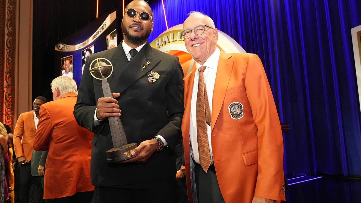 Dwóch mężczyzn pozuje do zdjęcia z trofeum. Po lewej stoi wysoki mężczyzna w czarnym garniturze, okularach przeciwsłonecznych i z zawieszonym na szyi trofeum. Po jego prawej stoi uśmiechnięty starszy mężczyzna w pomarańczowej marynarce.