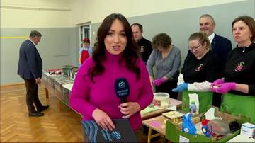 Kobieta z mikrofonem w r&oacute;żowym swetrze, z tyłu grupa os&oacute;b stojąca przy stołach z produktami.