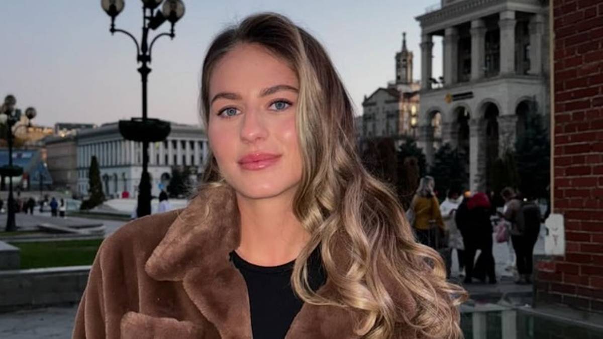 Portret kobiety z długimi blond włosami, ubranej w brązowe futro, stojącej na tle miejskiego krajobrazu z budynkami i latarniami.