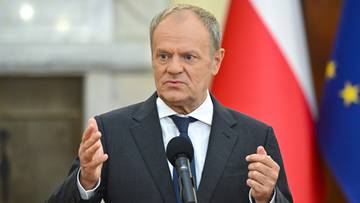 Premier Donald Tusk przemawia z m&oacute;wnicy, z flagą Polski i Unii Europejskiej w tle.