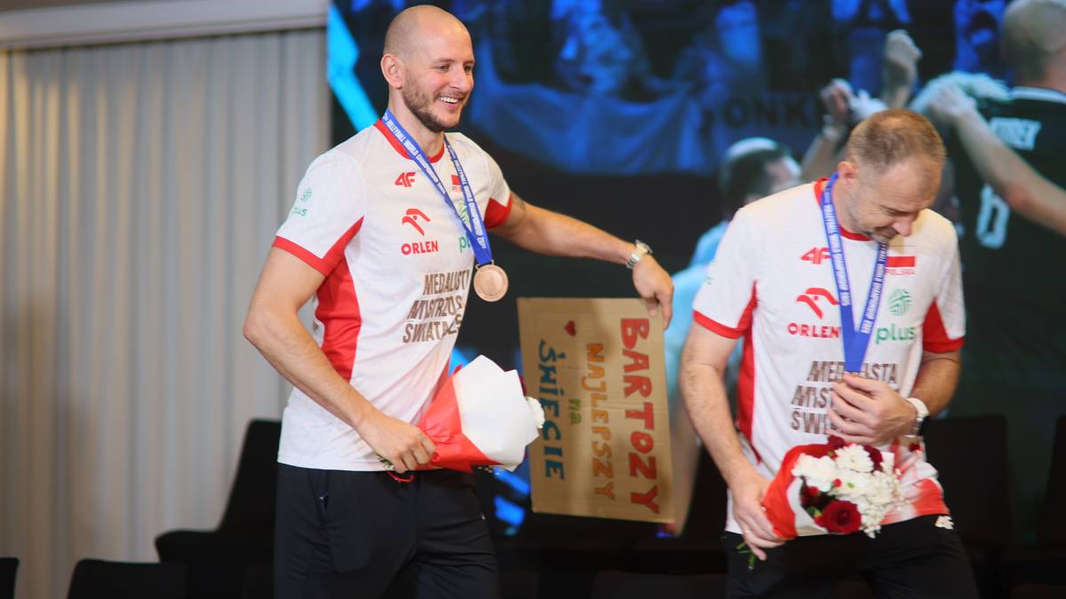 Dwóch mężczyzn w biało-czerwonych strojach sportowych z medalami na szyjach. Jeden z nich, z medalem na szyi i w dłoni, uśmiecha się. Drugi, również z medalem, trzyma w rękach bukiet kwiatów.