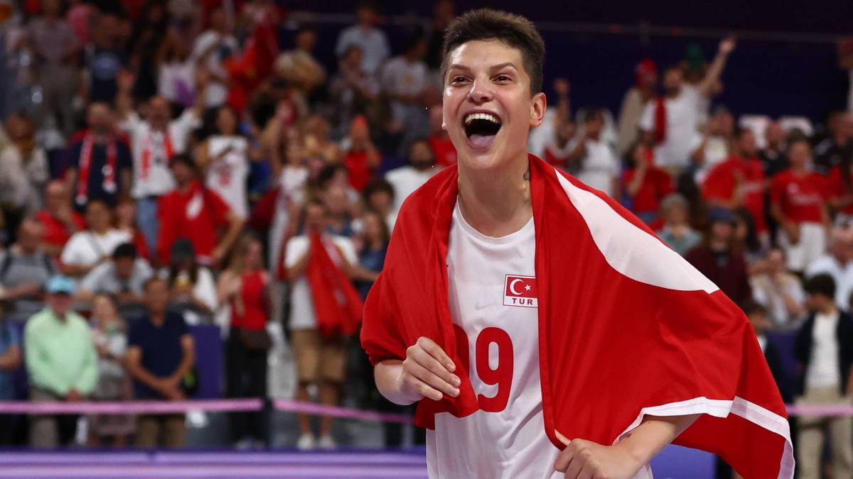 Siatkarka z Turcji triumfalnie unosi ręce, okryta turecką flagą. Widzowie na trybunach świętują w tle.