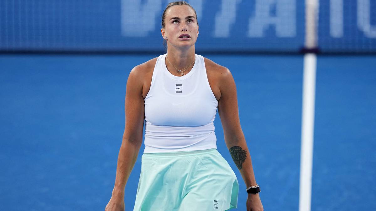 Białoruska tenisistka Aryna Sabalenka w białej koszulce sportowej i jasnoniebieskiej spódniczce stoi na korcie tenisowym, patrząc w górę.