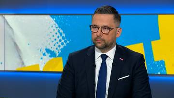 Mężczyzna w okularach i granatowym garniturze, ubrany w białą koszulę i niebieski krawat, siedzi w studiu telewizyjnym.