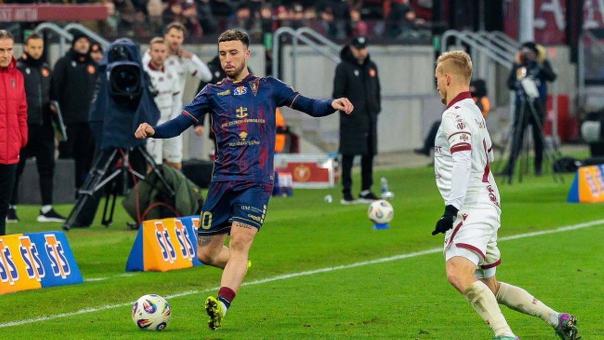 Piłkarz w granatowo-czerwonym stroju prowadzi piłkę, a przeciwnik w białym stroju próbuje go zatrzymać.