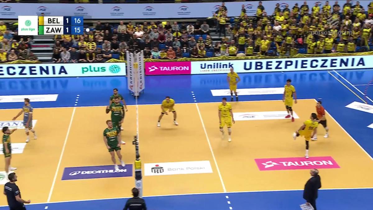 Gracze w żółtych i zielonych strojach grają w siatkówkę na boisku. Widoczni są kibice na trybunach.