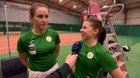 Dwie młode kobiety w zielonych koszulkach sportowych stoją obok siebie, jedna z nich trzyma mikrofon i rozmawia z drugą.