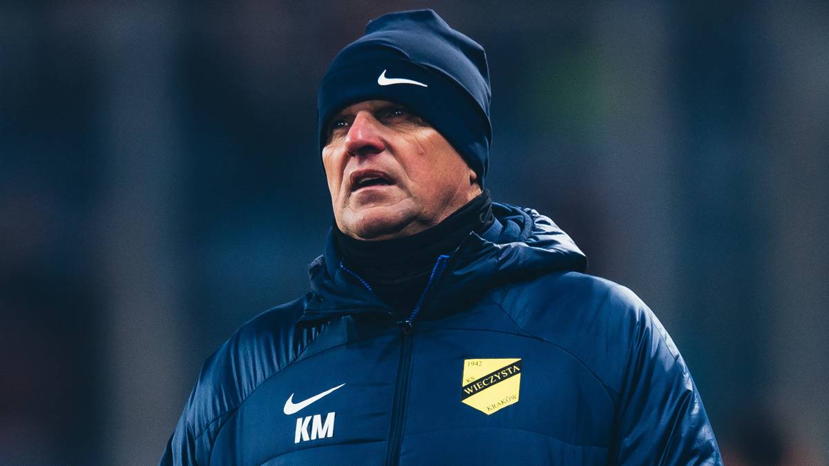 Trener piłkarski w zimowej kurtce i czapce, stojący na tle rozmytego stadionu.