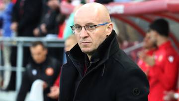 Trener w okularach i czarnym płaszczu podczas meczu piłki nożnej.