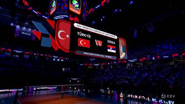 Widok na halę sportową podczas meczu siatkówki Turcja - Serbia, z widoczną podwieszaną tablicą wyników.