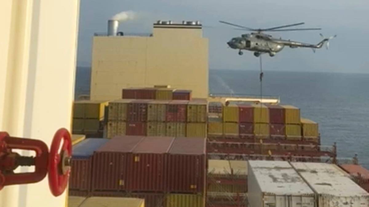 Helikopter zawisa nad statkiem towarowym załadowanym kontenerami, podczas gdy na pierwszym planie widoczna jest część instalacji przemysłowej statku.