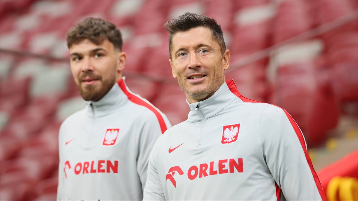 Dw&oacute;ch piłkarzy reprezentacji Polski w szarych strojach z czerwonymi elementami i godłem Polski, stojących przed czerwonymi trybunami stadionu.