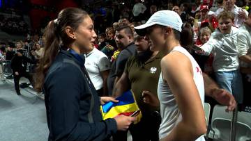 Tenisistka w ciemnej kurtce wręcza flagę innej tenisistce w białym stroju sportowym, na tle tłumu kibiców.