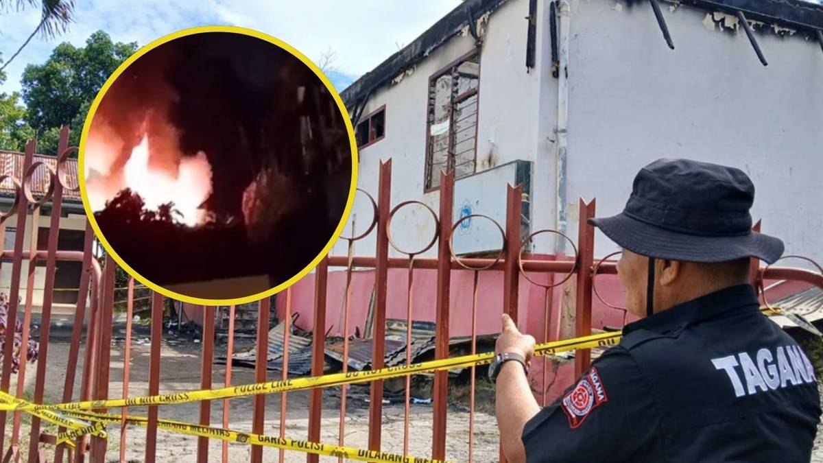 Funkcjonariusz policji w mundurze z napisem "TAGANA" na plecach wskazuje palcem w kierunku płonącego budynku, za metalowym ogrodzeniem z ż&oacute;łto-czarną taśmą policyjną.