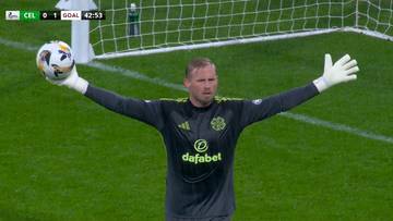 Bramkarz w czarnej koszulce z zielonym logo i białymi rękawicami bramkarskimi podnosi ręce na boki, trzymając piłkę w lewej dłoni. Na ekranie widoczny jest wynik meczu.