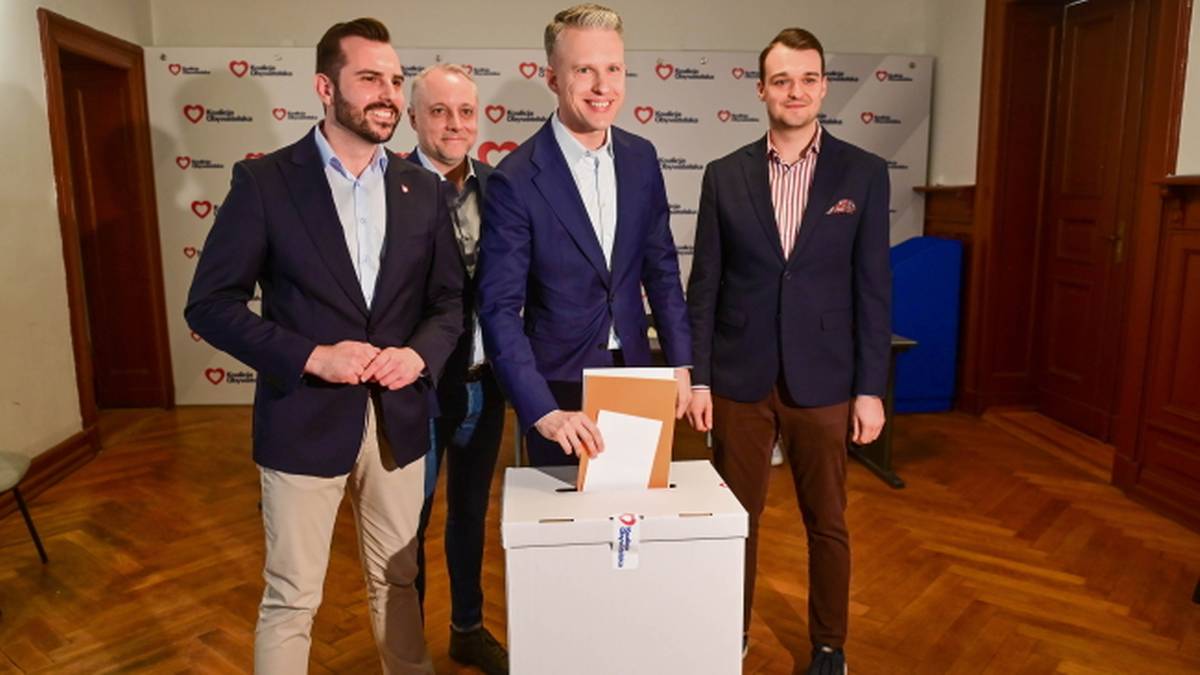 Czterech mężczyzn stoi przed białą urną wyborczą z logo Koalicji Obywatelskiej, z kt&oacute;rej jeden z nich wyjmuje kartę do głosowania.