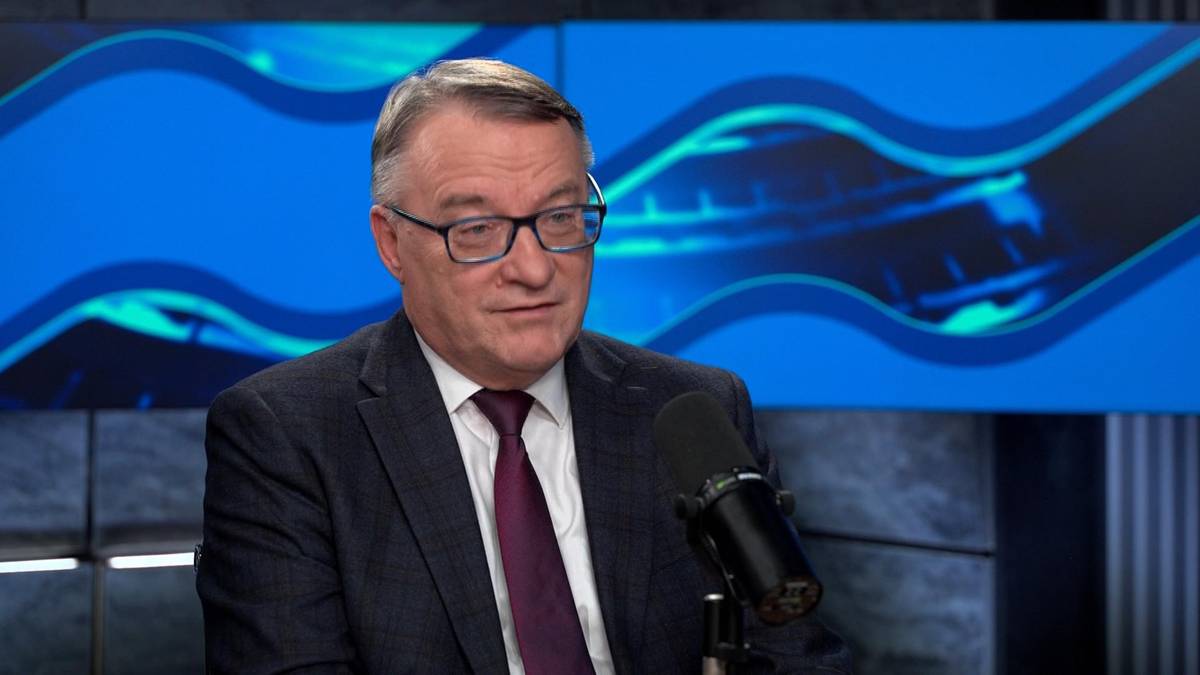 Marek Biernacki w okularach i garniturze siedzi przed mikrofonem na tle niebieskiego ekranu.