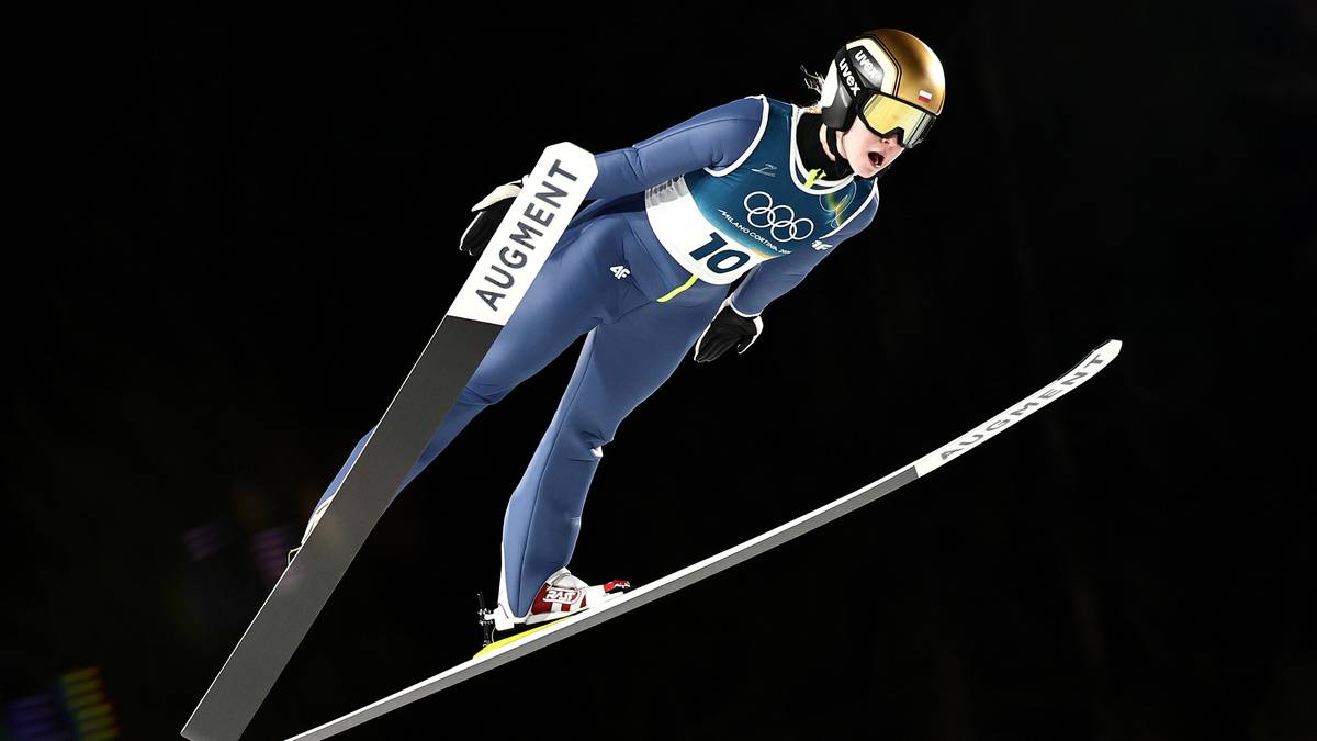 Skoczkini narciarska w powietrzu podczas zawodów, z nartami wyciągniętymi do przodu, ubrana w niebieski kombinezon z numerem 10.