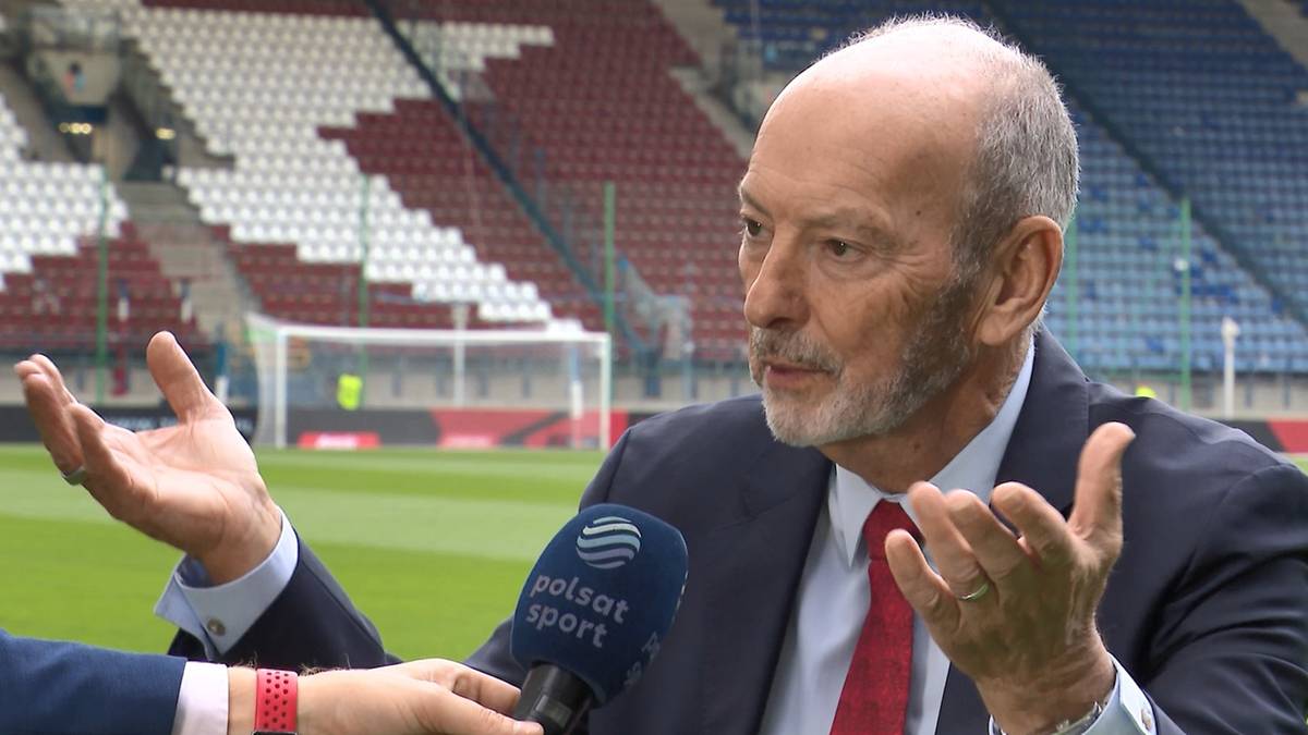 Peter Moore z siwą brodą, ubrany w ciemny garnitur i czerwoną koszulę, udziela wywiadu na tle stadionu piłkarskiego.