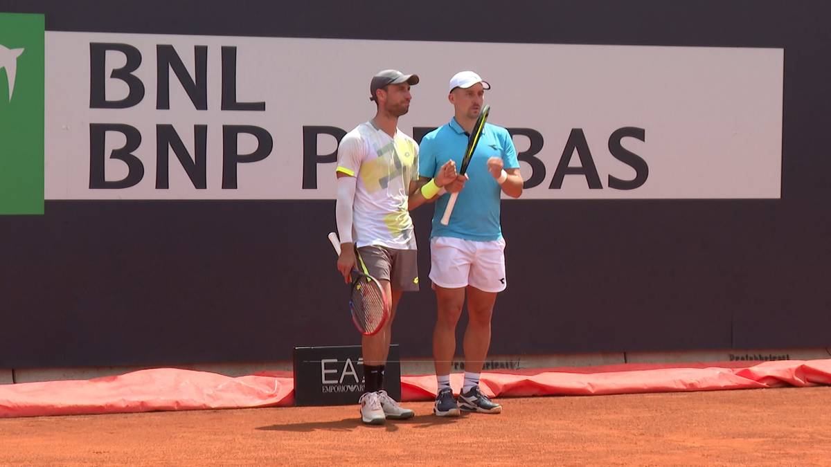 Dwóch mężczyzn w strojach tenisowych stoi na korcie ziemnym, trzymając rakiety. W tle znajduje się duży baner z logo.