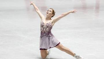 Łyżwiarka figurowa w fioletowej sukience z ozdobnymi wzorami na lodowisku, z uniesionymi ramionami i nogą w wykroku.