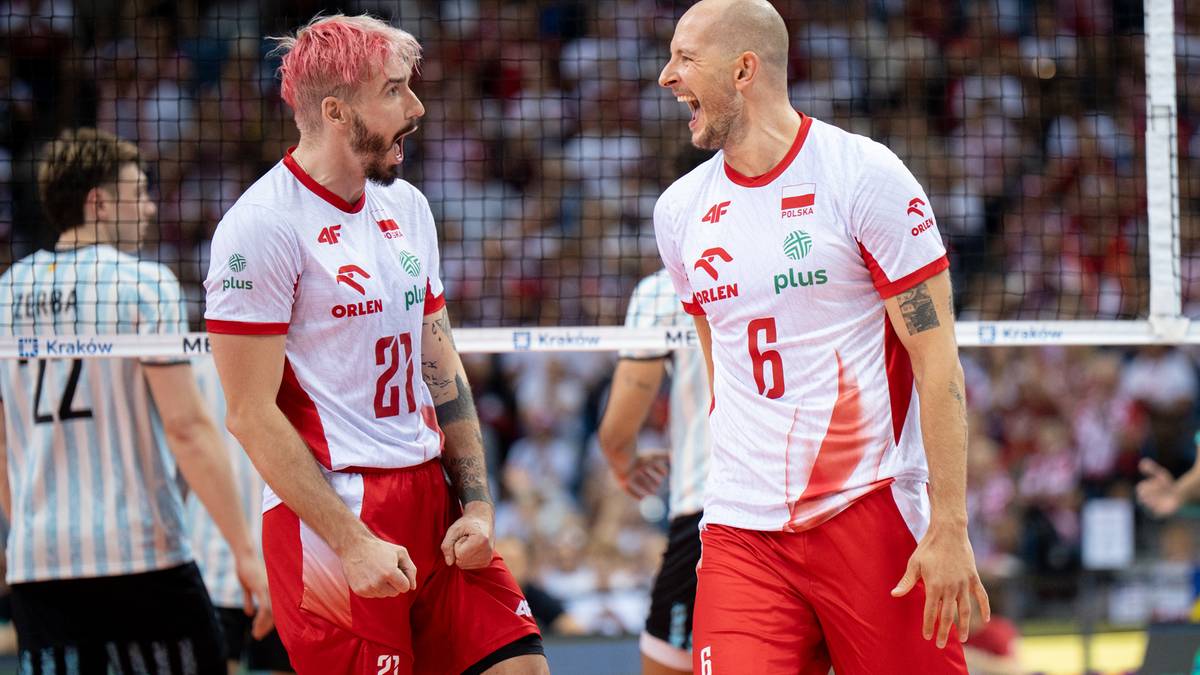 Dwóch zawodników reprezentacji Polski w siatkówce, ubranych w biało-czerwone stroje, podczas meczu. Jeden z zawodników ma krótkie, różowe włosy, drugi jest łysy. Obaj są w trakcie rozmowy i patrzą na siebie z intensywnością.