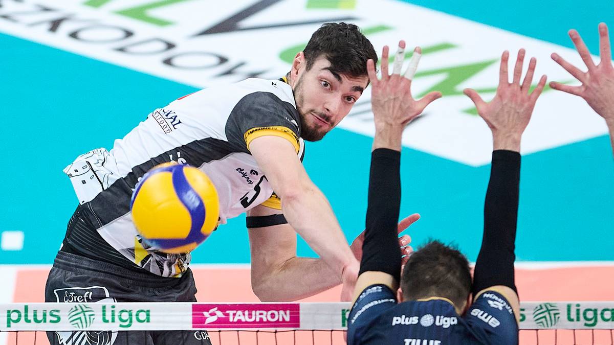 Siatkarz w białej koszulce z czarnymi elementami podaje piłkę, a po drugiej stronie siatki widać dłonie innego zawodnika pr&oacute;bującego ją zablokować.