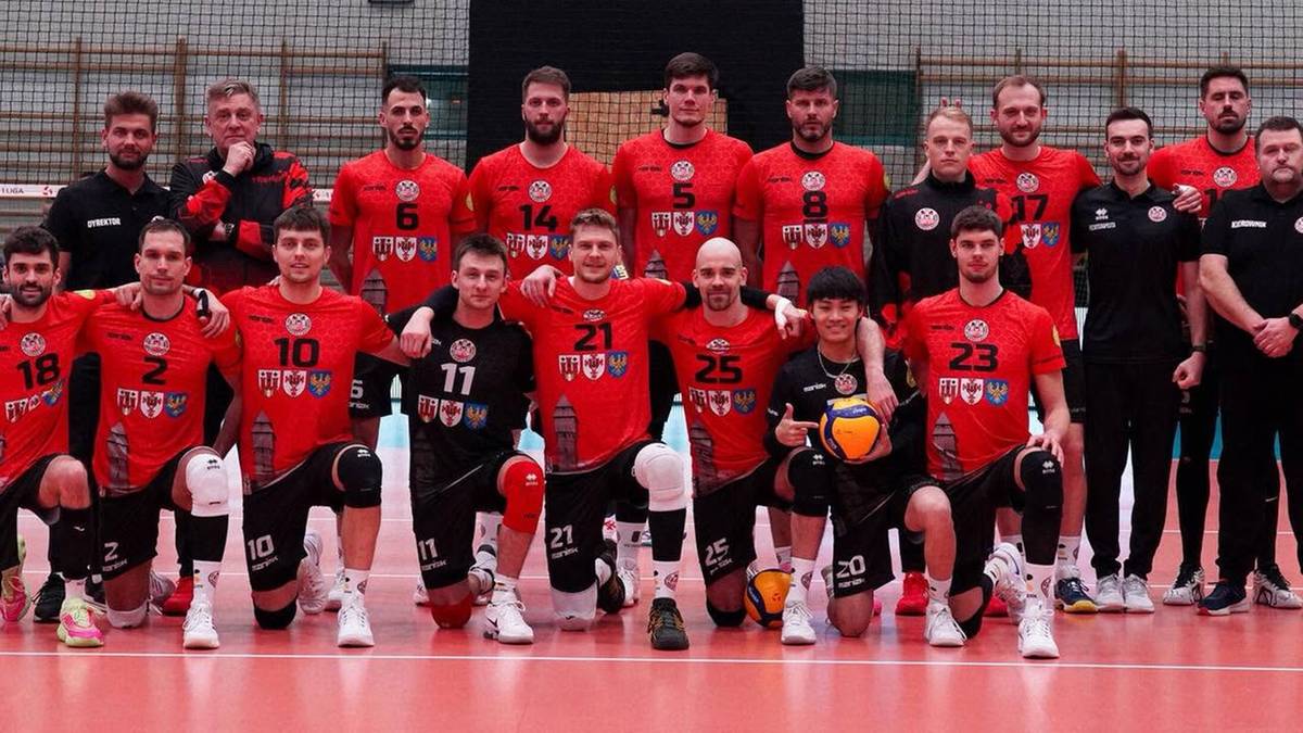 Drużyna siatkarska w czerwonych strojach pozuje do zdjęcia grupowego w hali sportowej.
