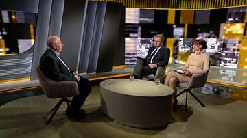 Dw&oacute;ch mężczyzn i kobieta siedzą na krzesłach wok&oacute;ł owalnego stołu w studiu telewizyjnym. Po lewej siedzi starszy mężczyzna w ciemnej marynarce i spodniach. Pośrodku siedzi mężczyzna w garniturze. Po prawej siedzi kobieta w jasnej sukience.