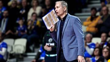 Trener drużyny sportowej w niebieskim garniturze trzymający kartkę z notatkami, wskazujący palcem i przemawiający do drużyny.
