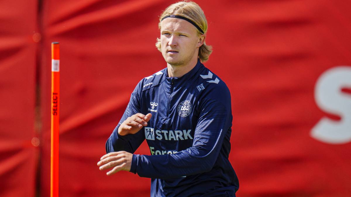 Piłkarz w niebieskiej bluzie treningowej z opaską na głowie, na tle czerwonego ogrodzenia.