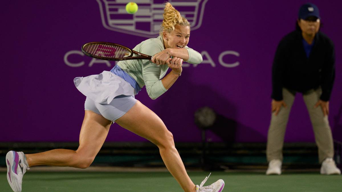 Tenisistka w akcji, uderza piłkę rakietą, w tle stoi osoba w ciemnej kurtce.