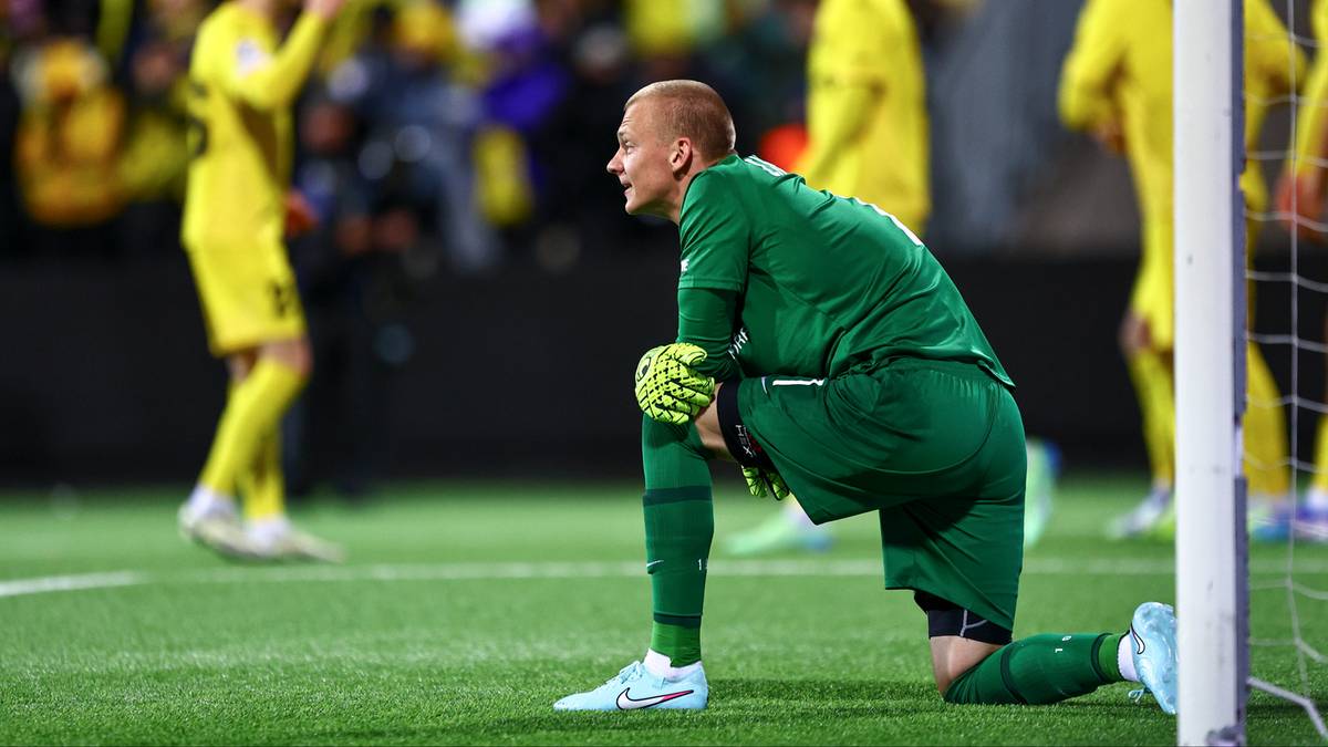 Bramkarz w zielonym stroju klęczy na murawie, patrząc w bok.