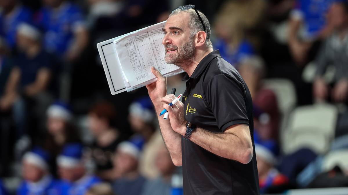 Mężczyzna w czarnej koszulce z żółtym logo, z wąsami i brodą, trzyma notes z notatkami i niebieski pisak. Ma na głowie opaskę.