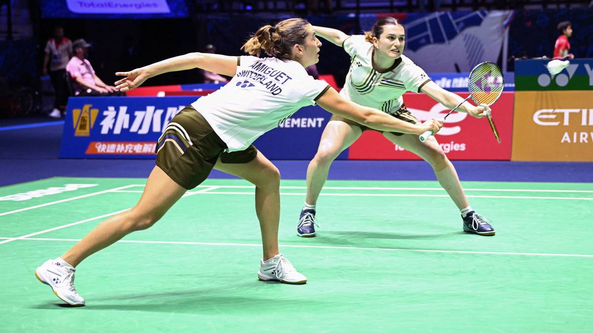 Dwie sportsmenki grające w badmintona. Jedna z nich, w białej koszulce, odbija piłeczkę rakietą, a druga, w zielono-białej koszulce, stara się ją zablokować.