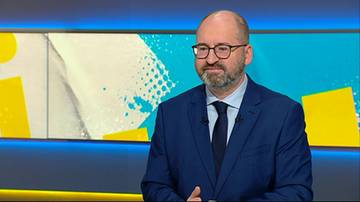 Mężczyzna w średnim wieku, ubrany w ciemnogranatowy garnitur, białą koszulę i czarny krawat, noszący okulary, z zarostem na twarzy, siedzi przed panelami z ż&oacute;łtymi i białymi elementami graficznymi na niebieskim tle.