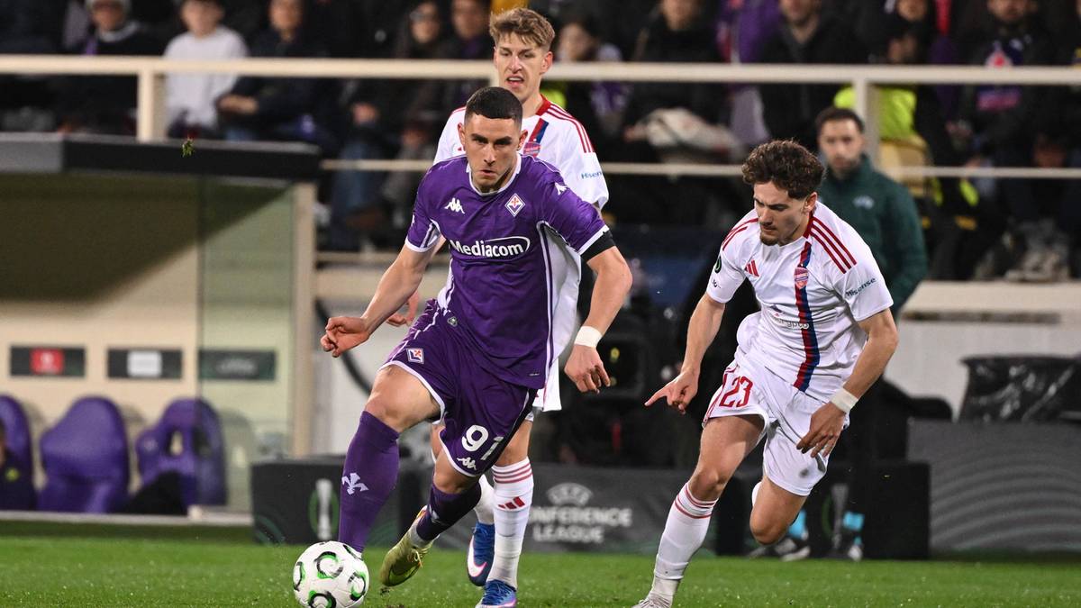 Dw&oacute;ch piłkarzy na boisku piłkarskim, jeden w fioletowej koszulce Fiorentiny z piłką przy nodze, drugi w białej koszulce Rakowa Częstochowa pr&oacute;bujący go zatrzymać.