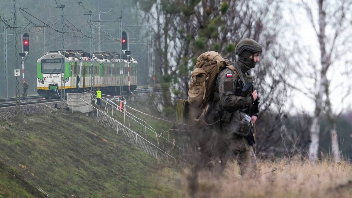 Żołnierz w pełnym umundurowaniu z plecakiem na tle torów kolejowych z pociągiem w tle.