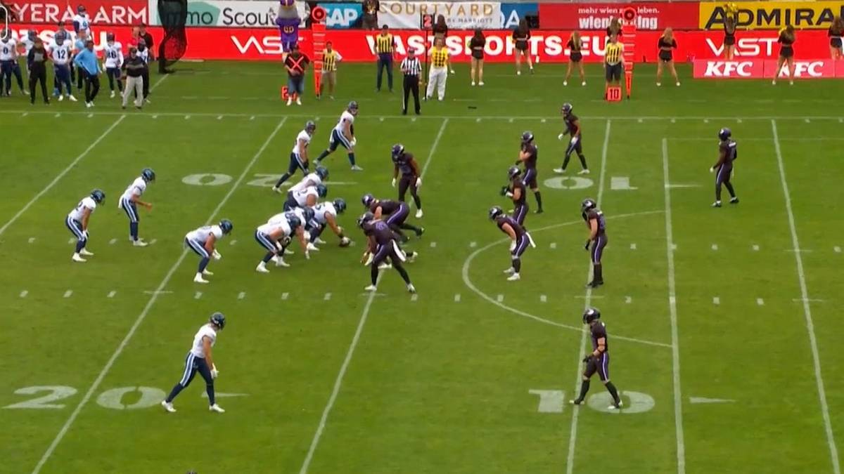 Gracze futbolu amerykańskiego w strojach fioletowo-czarnych i białych na zielonym boisku podczas meczu.