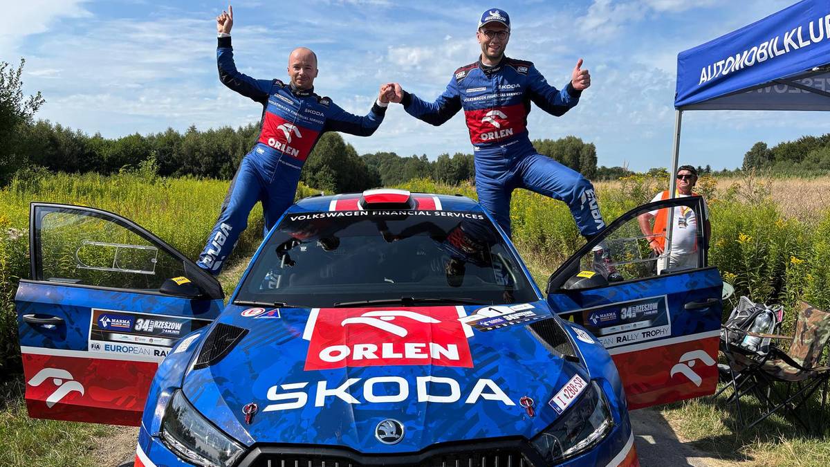 Dwóch mężczyzn w kombinezonach rajdowych stoi na masce samochodu Skoda podczas rajdu w Rzeszowie.