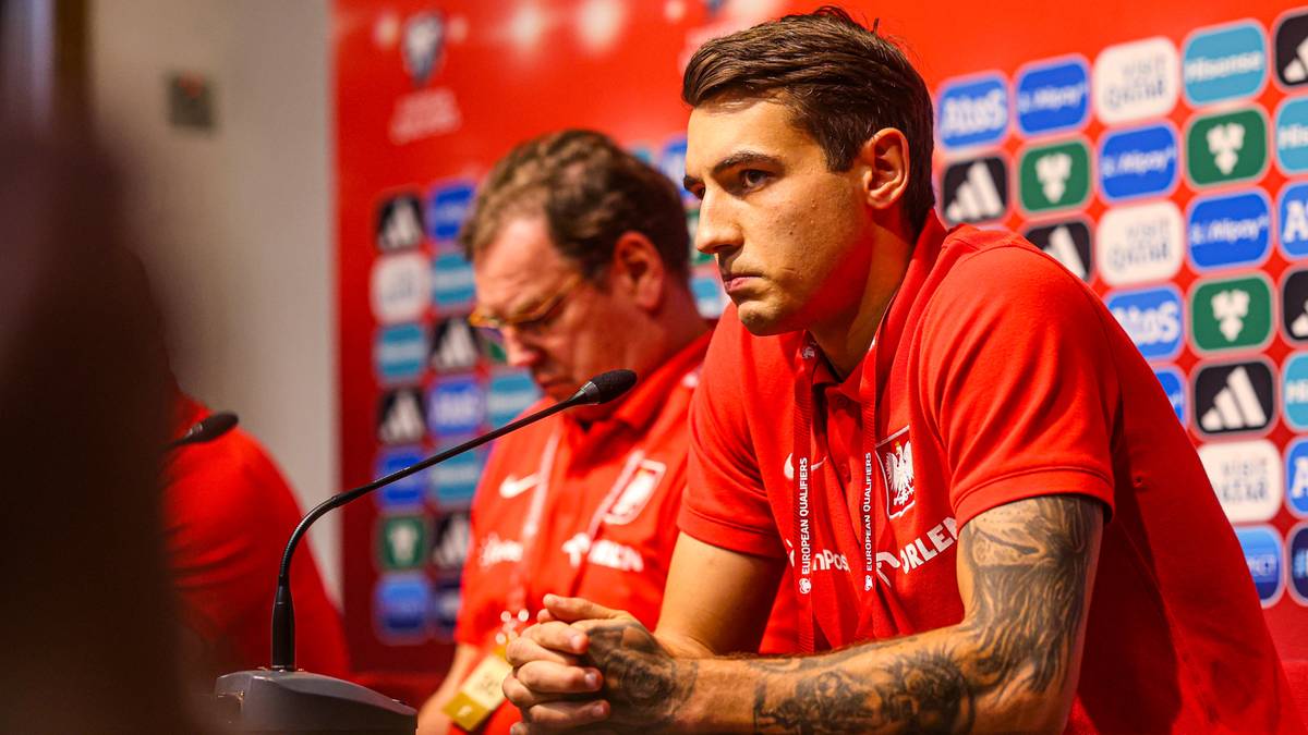 Dw&oacute;ch mężczyzn w czerwonych koszulkach z logo reprezentacji Polski, siedzi przed mikrofonem na tle ścianki z logotypami sponsor&oacute;w.