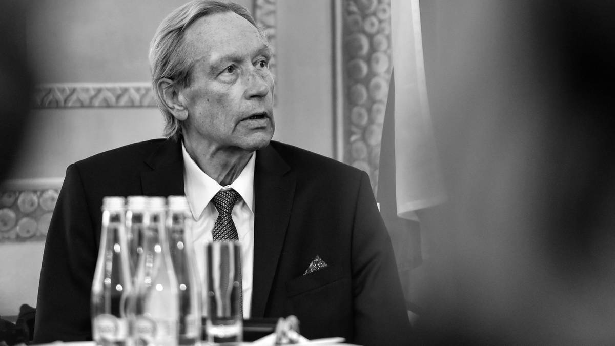 Portret starszego mężczyzny w garniturze i krawacie, siedzącego przy stole z butelkami wody i szklankami. Jest w kadrze od pasa w górę, patrzy w bok z poważnym wyrazem twarzy.