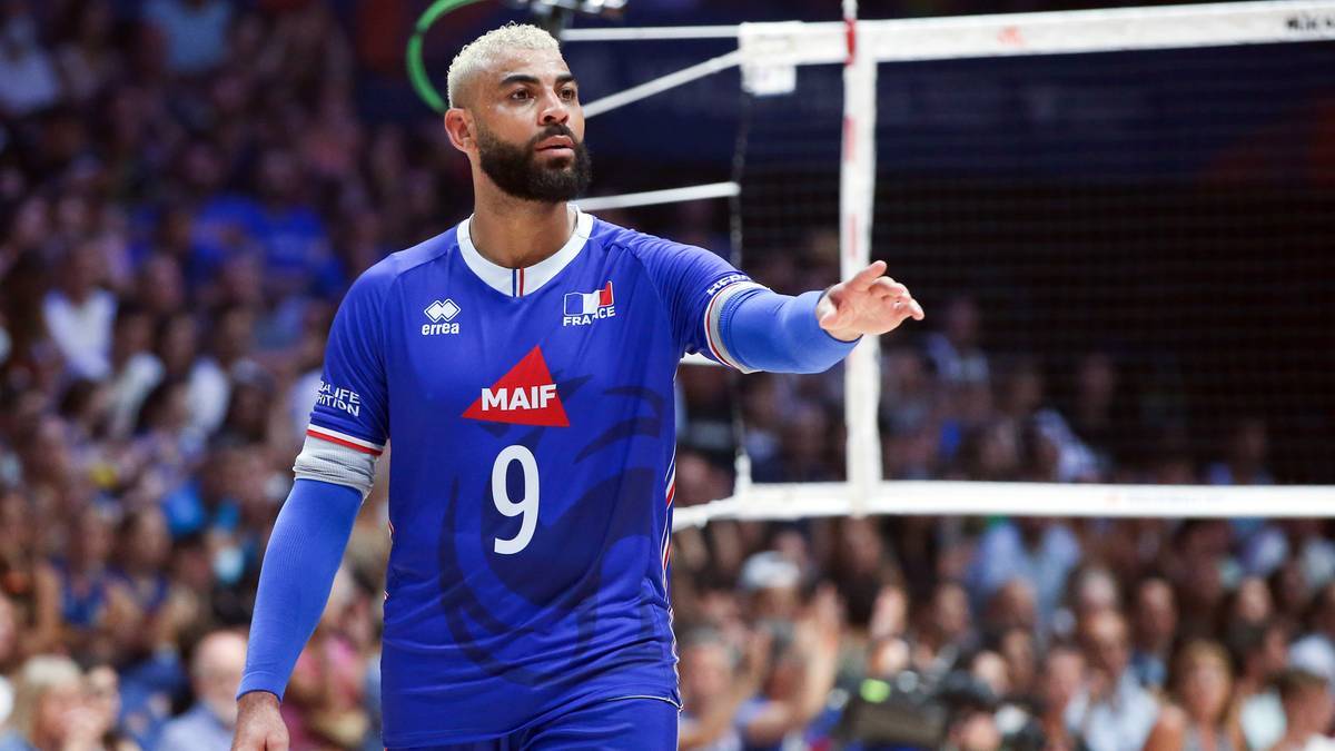 Siatkarz w niebieskiej koszulce reprezentacji Francji z numerem 9, z krótkimi blond włosami i brodą, na tle siatki i publiczności.