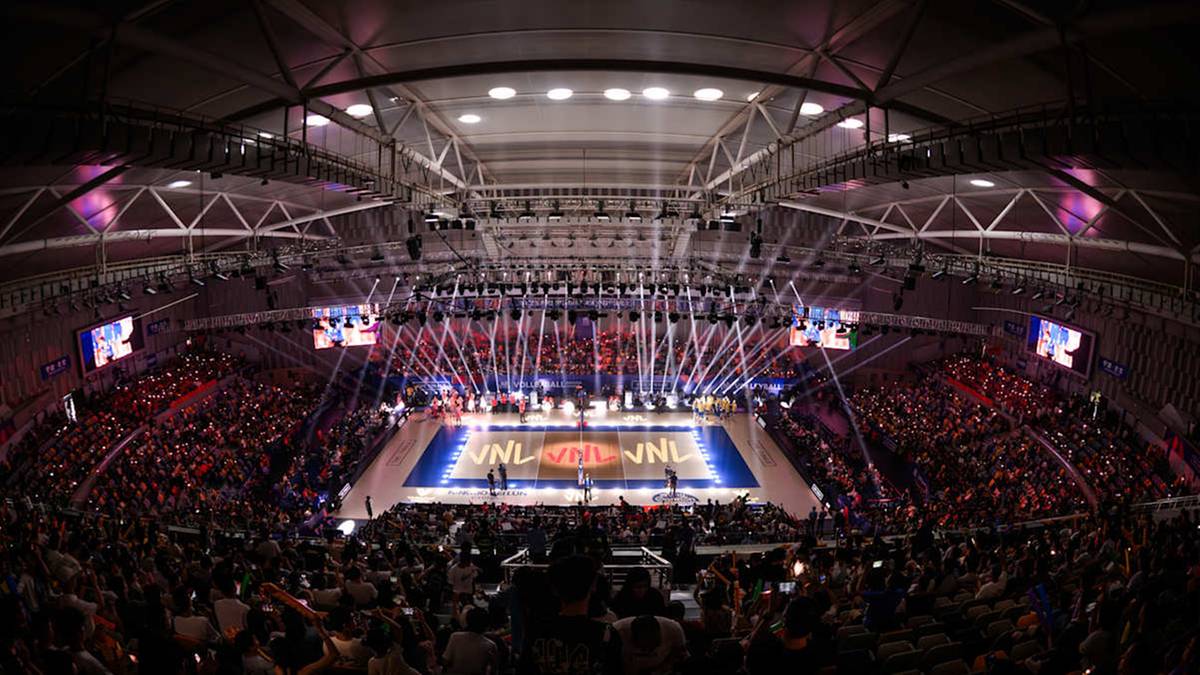 Rozciągnięty obraz hali sportowej z wypełnioną po brzegi widownią, oświetloną sceną na boisku do siatkówki.
