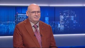 Mężczyzna w okularach i brązowej marynarce, stojący przed niebieskim ekranem z widokiem nocnego miasta.