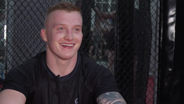 Portret uśmiechniętego zawodnika MMA w czarnej koszulce przed klatką ringową.