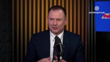 Mężczyzna w ciemnym garniturze i białej koszuli z czarnym krawatem rozmawia, patrząc w stronę kamery. Przed nim znajduje się mikrofon. Za nim znajduje się tło w ciemnych odcieniach z pionowymi pasami i logo "polsatsport.pl".